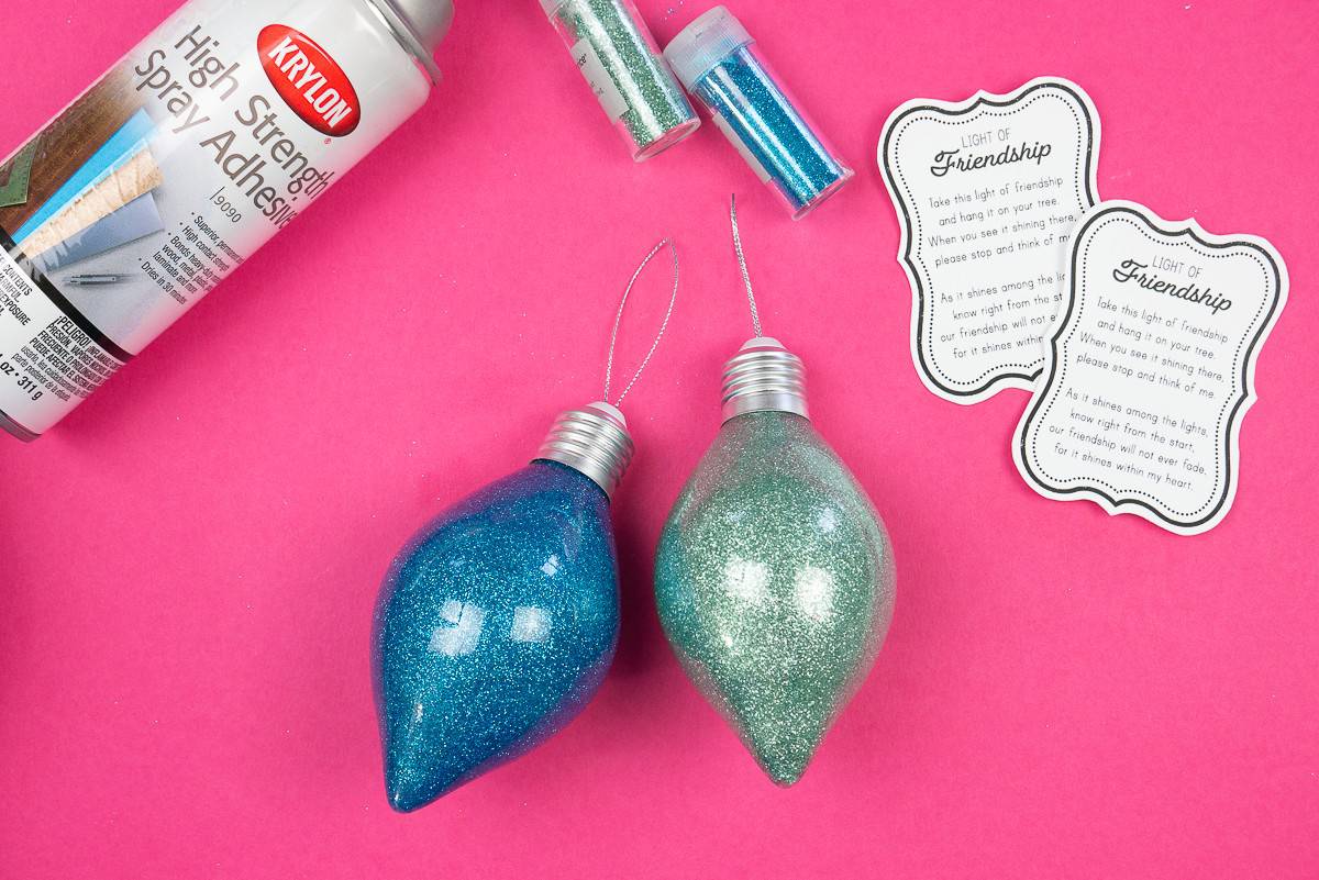 Two glittered ornaments on a pink background with supplies.